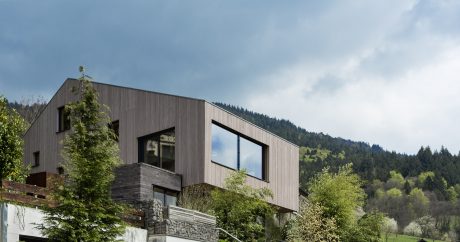 A modern, multi-story building with wood and stone exterior, surrounded by lush greenery.