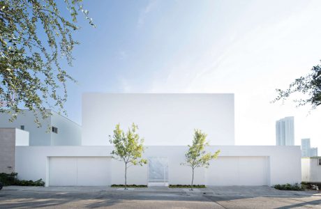 Sleek, modern white building with glass facade and trees in foreground against blue sky.