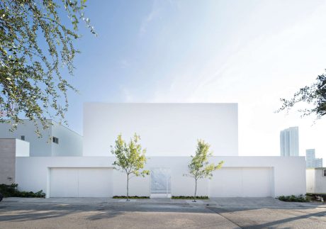 Sleek, modern white building with glass facade and trees in foreground against blue sky.