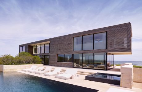 Sleek modern beachfront house with large glass walls, outdoor seating, and a pool.