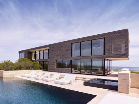 Sleek modern beachfront house with large glass walls, outdoor seating, and a pool.