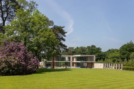 Modern two-story home with glass walls, surrounded by lush greenery and blooming trees.