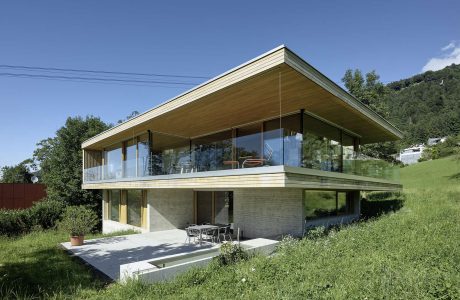 Sleek, modern house with expansive glass walls, wood-paneled roof, and grassy surroundings.