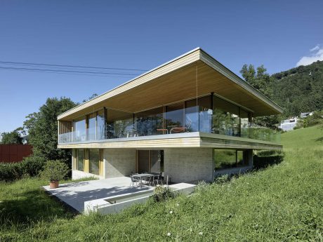 Sleek, modern house with expansive glass walls, wood-paneled roof, and grassy surroundings.