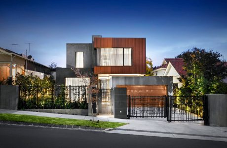 A modern residential building with a mix of concrete, wood, and metal accents, surrounded by lush greenery.