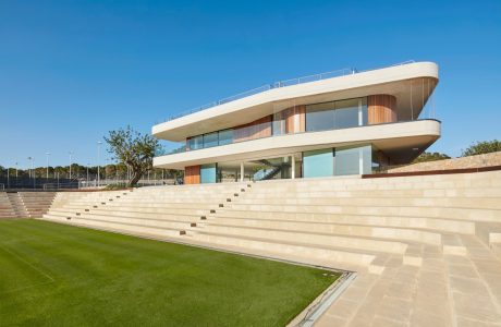 A modern, multi-level building with sleek, curved lines, expansive glass panels, and a grand staircase leading to a grassy field.