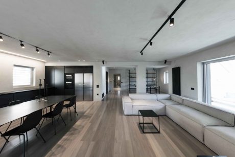 Spacious, minimalist living space with sleek black kitchen, wooden flooring, and track lighting.