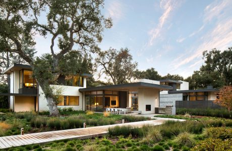 A modern, two-story home with large windows, surrounded by lush greenery and a wooden bridge.