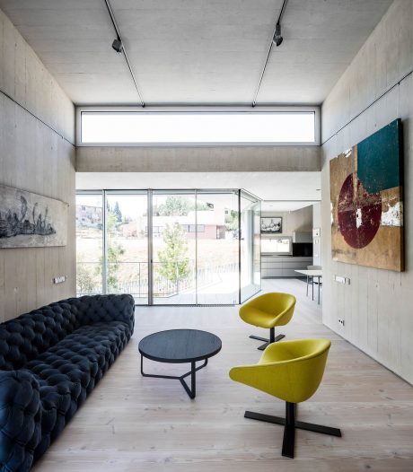 A modern, minimalist living space with concrete walls, large windows, and a mix of black and yellow furniture.