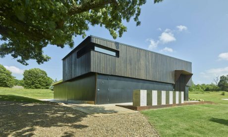 A modern, rectangular building with dark wooden siding and concrete pillars, situated in a grassy, wooded area.