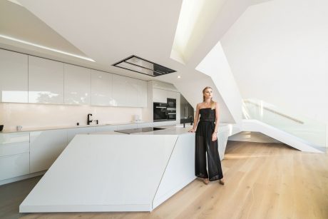 Modern, minimalist kitchen with white cabinets, dark countertop, and a sleek, stylish woman standing in the room.