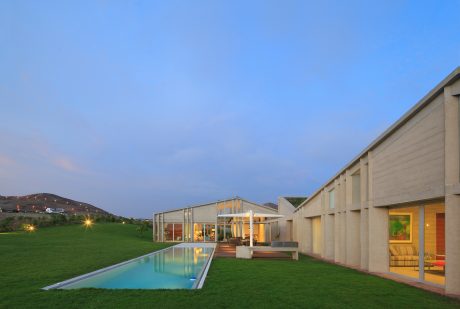 Modern, open-plan home with glass walls, pool, and lush green lawn against a blue sky.