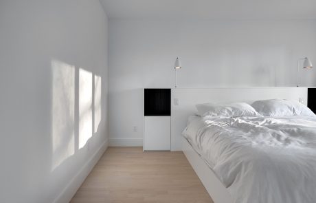 Minimalist bedroom with white walls, large windows, wooden floors, and simple black furniture.