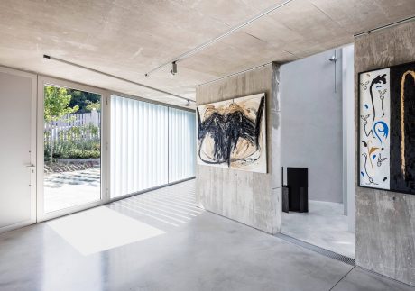 Contemporary open-plan interior with concrete walls, artwork, and large windows overlooking garden.