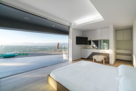 Spacious modern bedroom with floor-to-ceiling glass walls, minimalist furniture, and cityscape view.