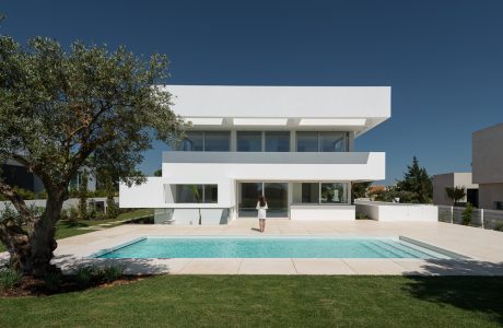 A sleek, modern white house with large windows, a pool, and a well-manicured lawn against a blue sky.