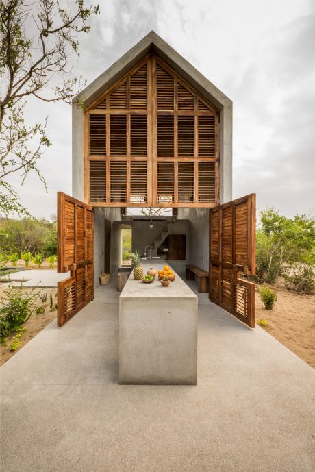 A modern architectural design featuring a wooden structure with shuttered windows, showcasing a concrete kitchen counter surrounded by tropical foliage.