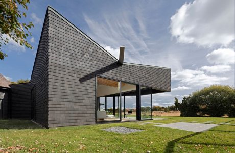 A modern, angular house with large windows, surrounded by a lush lawn and trees.