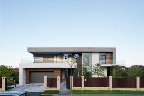 A modern two-story house with a concrete exterior, glass walls, and wooden accents. A dark wooden fence surrounds the property.