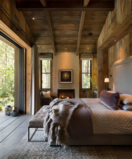 Rustic cabin interior with wooden beams, fireplace, and expansive windows overlooking nature.