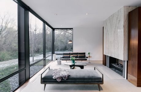 A modern, minimalist living room with large windows overlooking a wooded landscape.