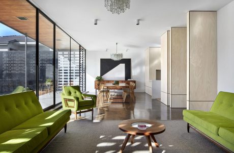 Bright, modern open-plan living space with green upholstered furniture, wood accents, and large glass windows.