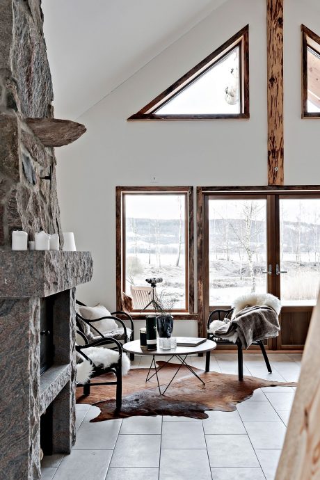 Cozy cabin interior with stone fireplace, wood accents, and large windows overlooking snowy landscape.