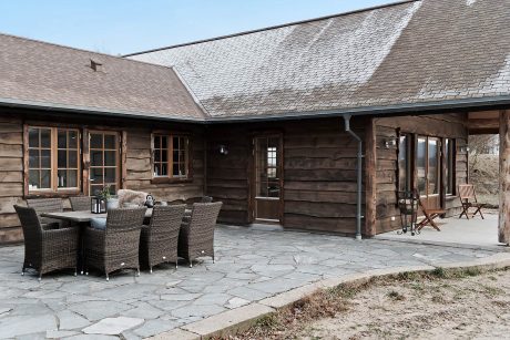 A rustic log cabin with a covered patio, wicker furniture, and a stone floor.