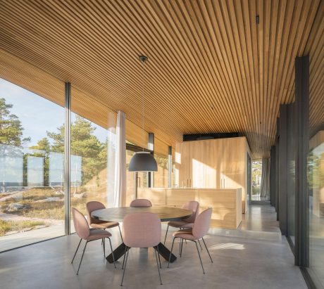 A modern dining room with a wooden slat ceiling, concrete floor, and large windows overlooking nature.