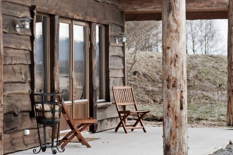 Rustic log cabin exterior with wooden chairs on a covered porch, surrounded by nature.