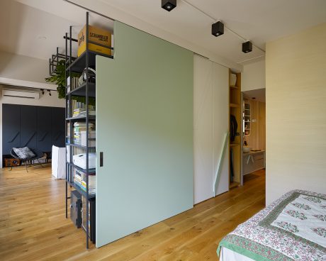 A modern, minimalist studio apartment with a green accent wall, shelving, and hardwood floors.