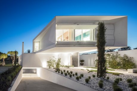 Modern, minimalist home with angled white facade, large windows, and lush landscaping.