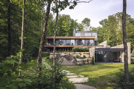 A modern, glass-walled house nestled among lush greenery with stone steps leading to the entrance.