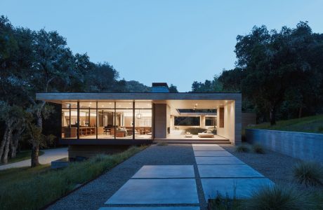 A modern, glass-walled house nestled among trees, with a sleek patio and reflecting pool.