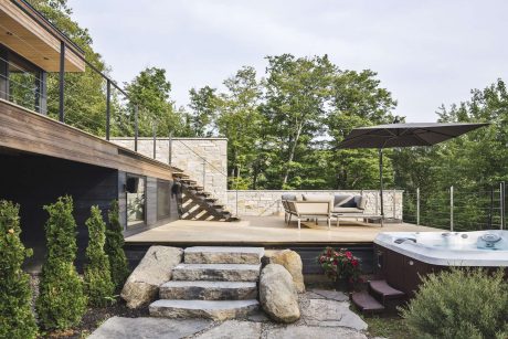 A modern outdoor living space with a large stone deck, stairs, and hot tub surrounded by lush greenery.