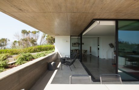 Spacious covered balcony with concrete ceiling and sliding glass doors, surrounded by lush greenery.