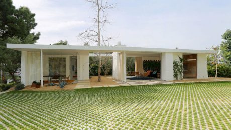 A mid-century modern house with a large covered porch, surrounded by a lush, patterned lawn.