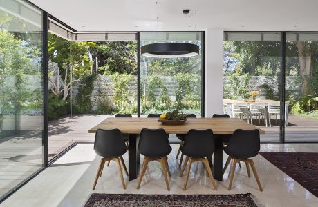 Modern open-plan dining area with a large wooden table, black chairs, and floor-to-ceiling windows overlooking a lush garden.