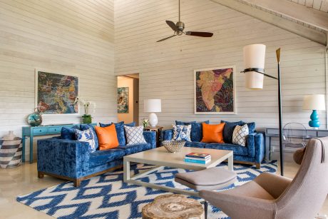 A cozy living room with a distinctive blue velvet sofa, vibrant artwork, and intricate patterned rug.