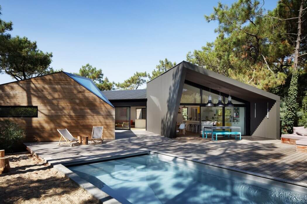 Modern wooden house with glass walls, pool, and outdoor furniture in lush forest setting.