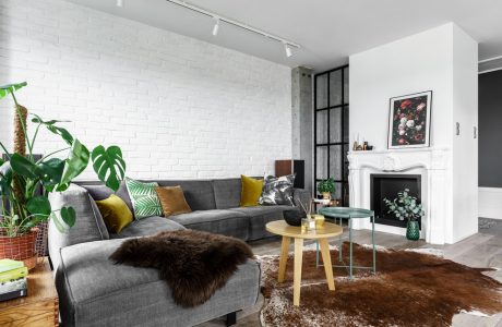 Cozy living room with gray couch, potted plants, fireplace, and furry rug on hardwood floor.