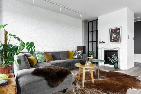 Cozy living room with gray couch, potted plants, fireplace, and furry rug on hardwood floor.