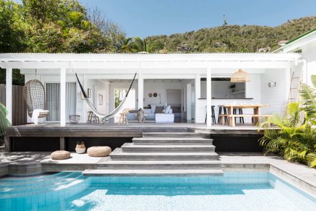 A modern, minimalist home with a pool, wooden deck, and tropical vegetation.