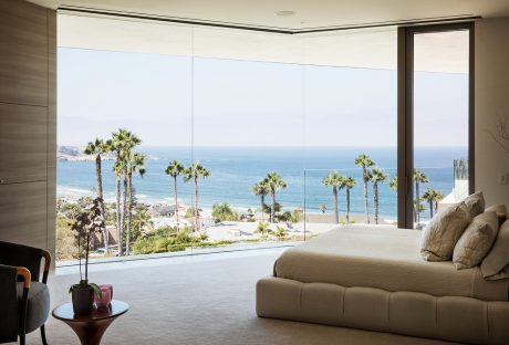 Spacious bedroom with floor-to-ceiling windows offering stunning ocean views and palm tree scenery.