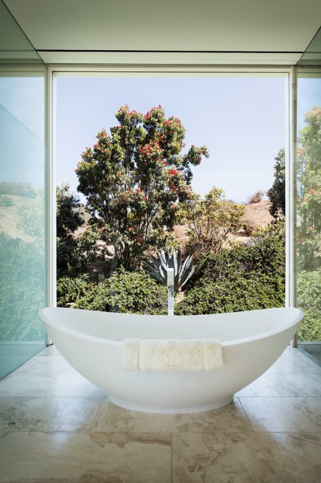 A stylish, freestanding white bathtub set against a lush outdoor landscape, framed by large glass windows.