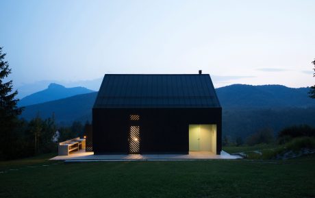 A modern, sleek black cabin nestled in a mountainous landscape, with a covered patio and large windows.