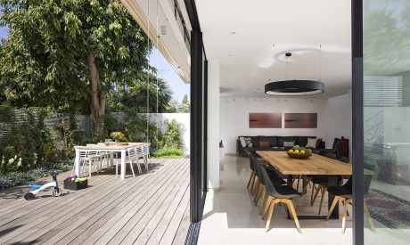 A modern open-concept living area with a wooden dining table, hanging light fixture, and glass wall overlooking lush greenery.