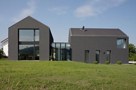 A modern, minimalist house with large glass windows and a slanted roof, surrounded by greenery.