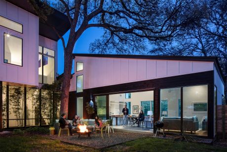 A modern glass-enclosed patio with a firepit, surrounded by a large oak tree and landscaping.