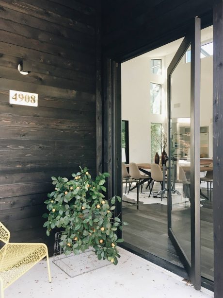 Rustic wooden exterior, sleek glass entryway, potted foliage, and modern furnishings inside.